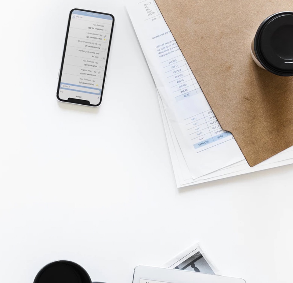 Birds eye view of a cell phone, to go coffee cup with a black plastic lid, and a brown folder with paperwork on a white background